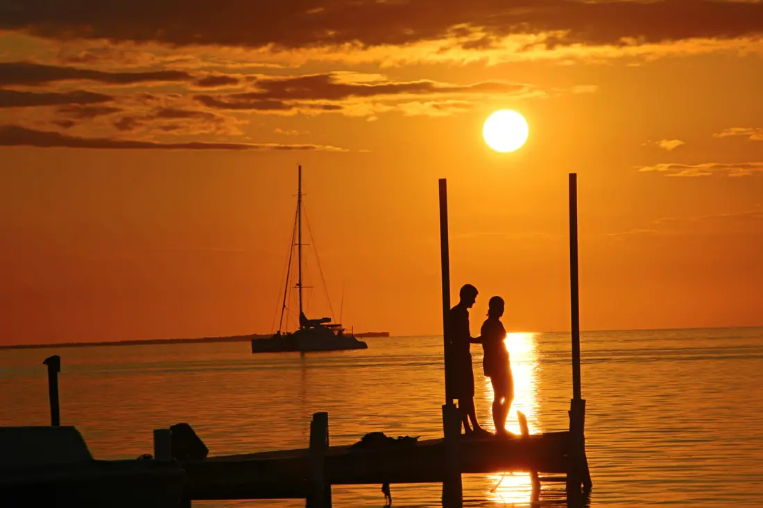 A couple silhouetted on a dock at sunset, a sailboat on the horizon
