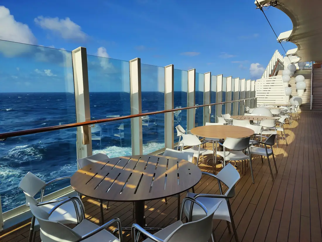 An open-air ship deck with tables beside a glass railing and the blue sea